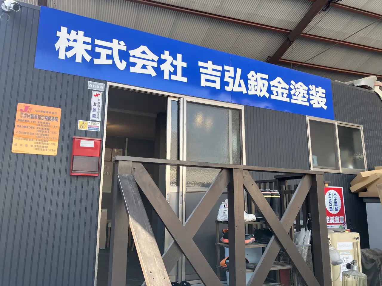 株式会社吉弘鈑金塗装の事務所外観。青い背景に白文字で「株式会社吉弘鈑金塗装」と大きく書かれた看板が入り口の上に掲げられている。