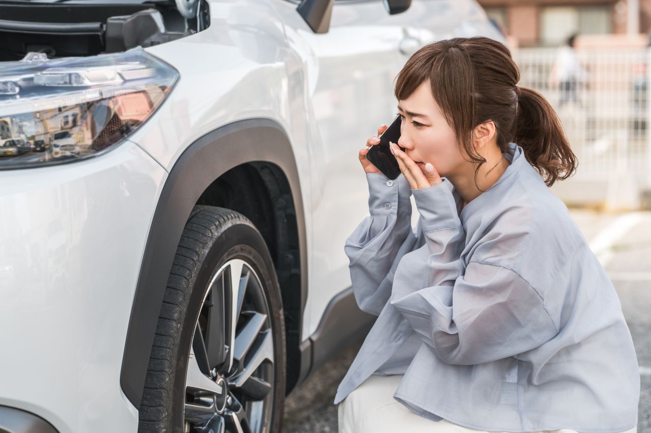 ボンネットを開けた白いSUVの横で、女性が不安そうな表情でスマートフォンを使い電話をしている